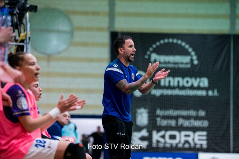 Coach in blue shirt claps and encourages a team on the sideline during a indoor sports game.