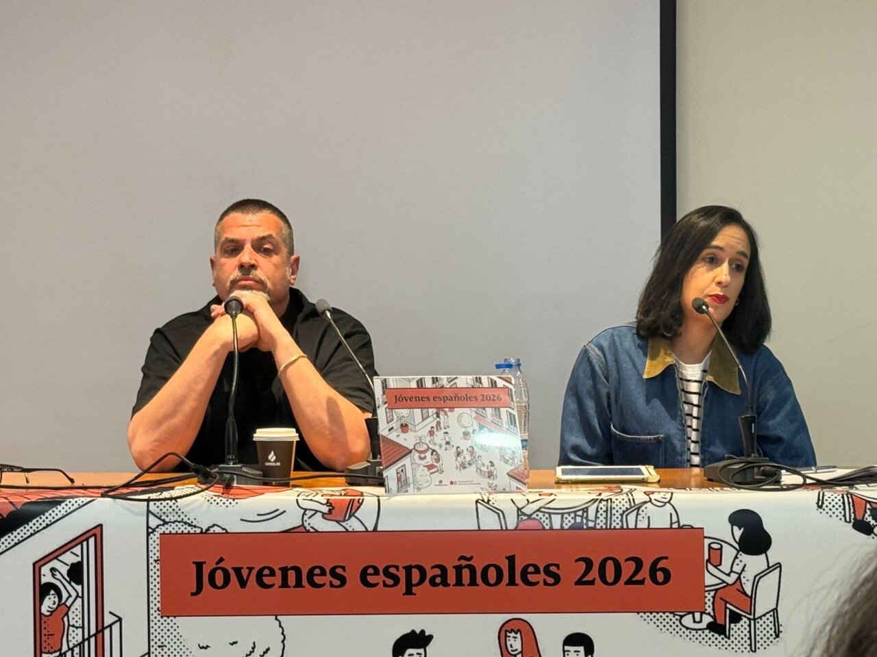 Two panelists sit at a long table with microphones, a coffee cup, and a sign reading 'Jóvenes españoles 2026' in front of them.