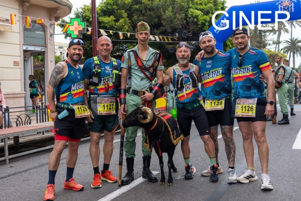 Six runners in blue race jerseys pose on a city street with a black ram wearing ceremonial gear between them, bib numbers visible on their shorts.