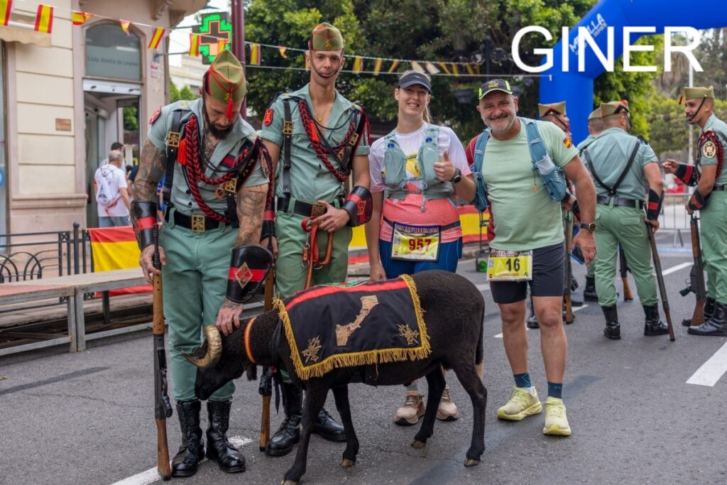 Group of people in green ceremonial uniforms and runners pose on a city street with a decorated black sheep at a race/event.