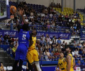 Blue-team player (#5, Heron) leaps to block a shot at the basket as a yellow-team defender confronts him; crowd watches from bleachers nearby.