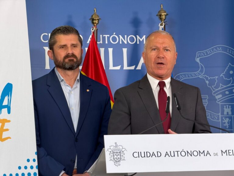 Two men in suits stand at a press podium, with a blue backdrop that reads Ciudad Autónoma de Melilla and a flag behind them.