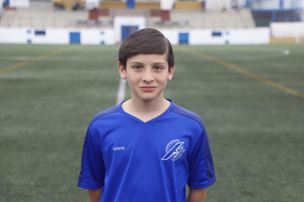 Boy in a blue soccer jersey stands on a grassy field with empty stadium stands in the background, looking at the camera.