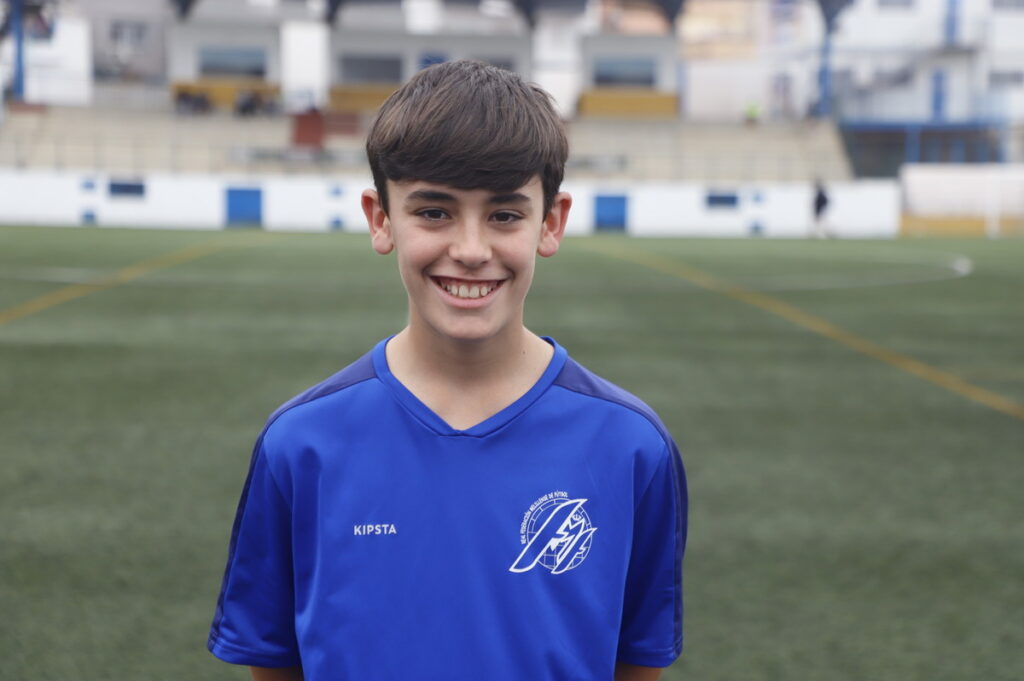 Smiling boy in a blue soccer jersey standing on a grassy field with stadium seating in the background