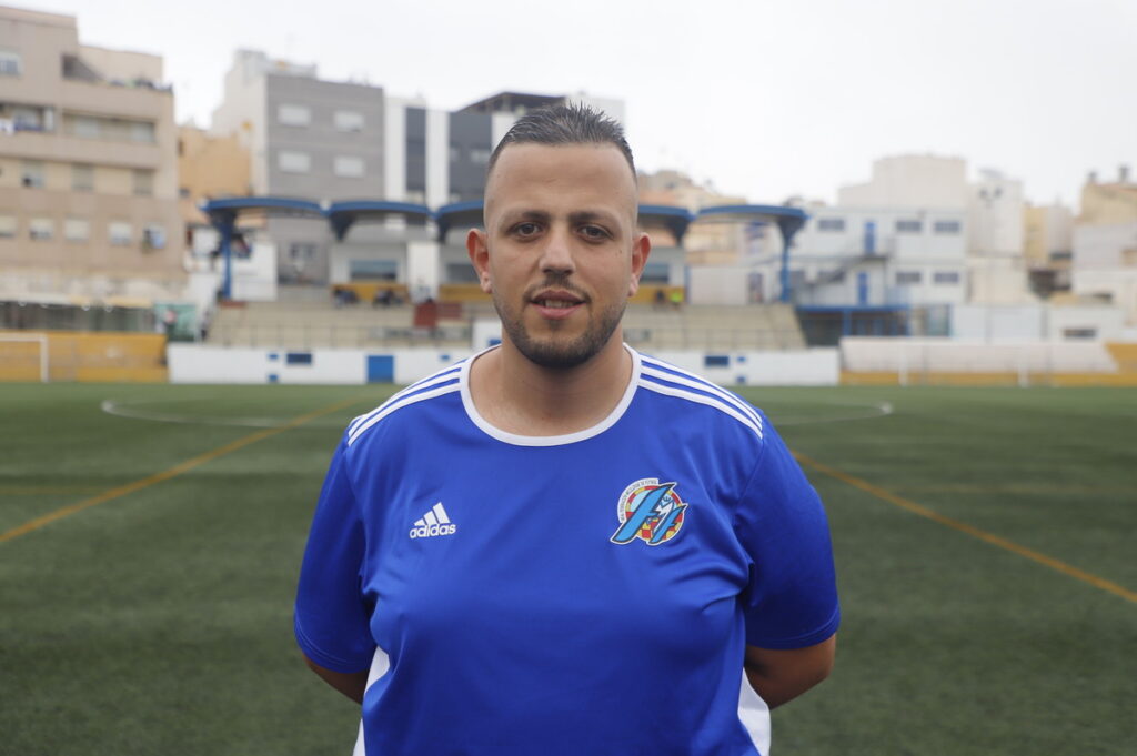 Portrait of a man in a blue soccer jersey standing on a soccer field with stadium stands in the background, facing the camera.