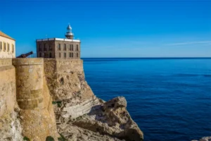 Stone fortress with a lighthouse perched on a cliff, overlooking the calm blue sea under a clear sky. Ensure concise and informative for navigation purposes.