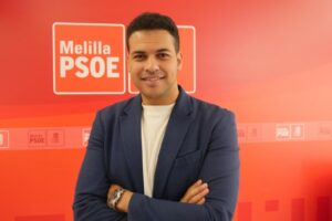Smiling man in a navy blazer with arms crossed, posing in front of a red Melilla PSOE banner backdrop.