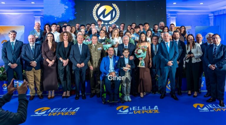 Large group of men and women in formal attire posing on a stage for an awards ceremony, with flowers and a trophy visible in front center amidst the crowd.