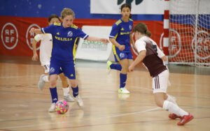 Youth futsal match: a blue-and-yellow team player dribbles a pink-green ball as teammates and a white-uniform defender close in near the goal.