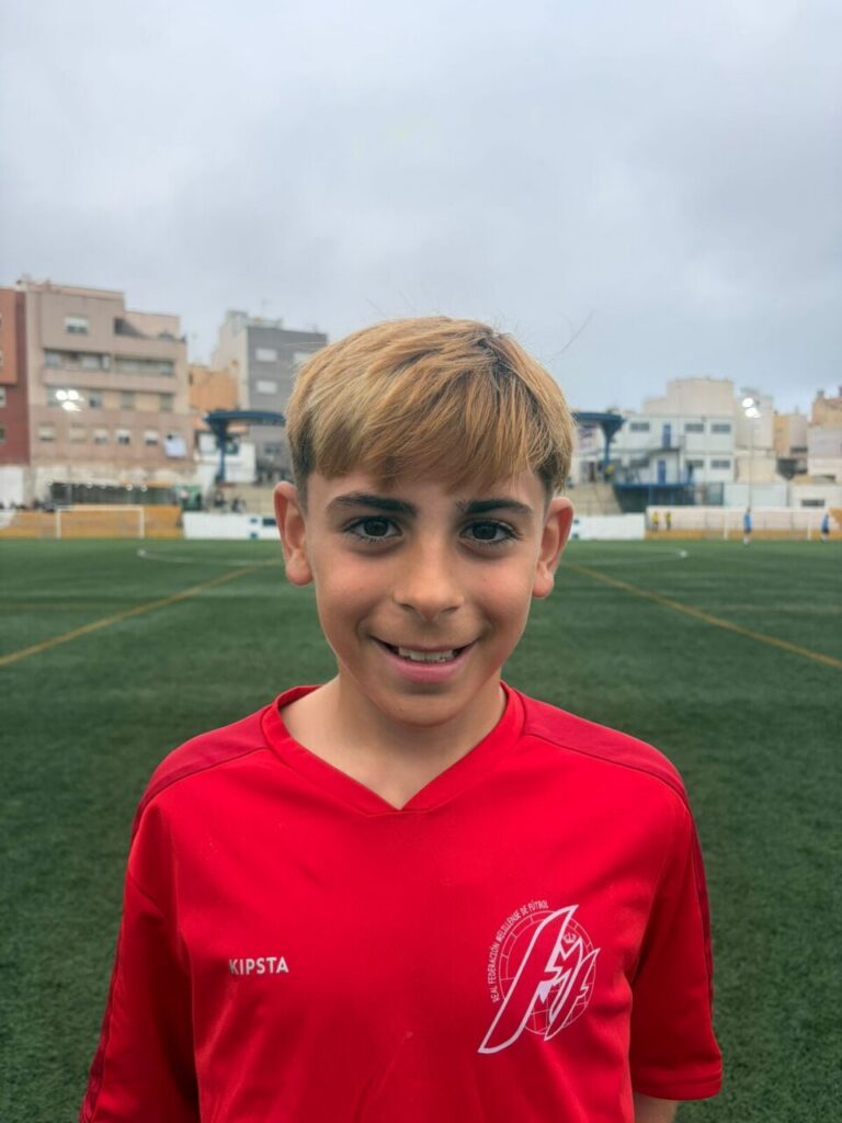 Boy in a red soccer jersey standing on a green outdoor field with buildings and an overcast sky in the background.