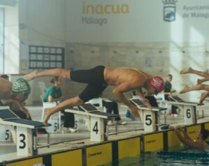 Swimmers dive from starting blocks at an indoor competition; lane numbers 3–6 visible with banners in the background.