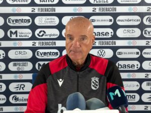 Bald man in a red and black jacket speaks at a press conference with multiple microphones and a sponsor backdrop.