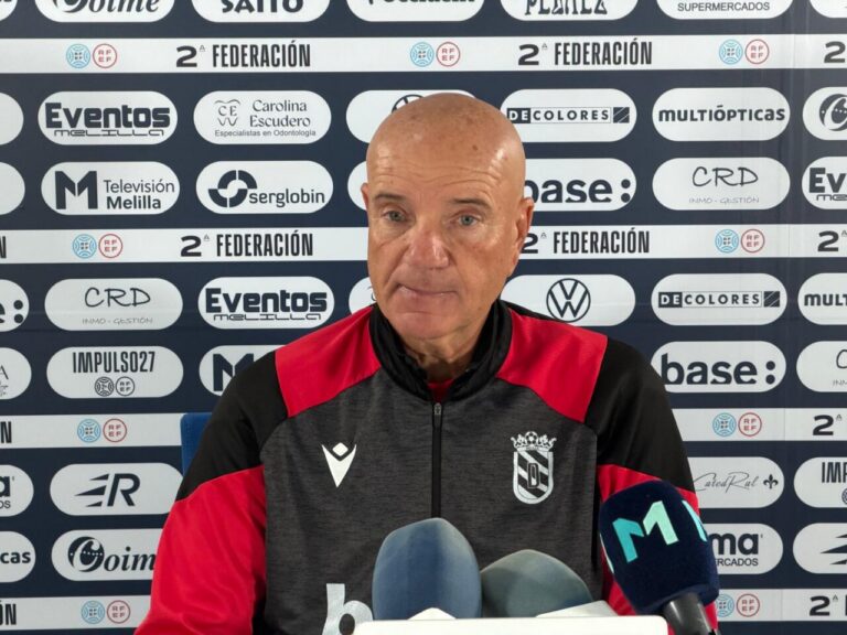 Bald man in a red and black jacket speaks at a press conference with multiple microphones and a sponsor backdrop.