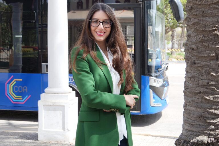 Mujer con blazer verde sonriendo frente a un autobús