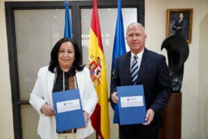 Two officials in business attire hold blue folders with an emblem, standing before Spanish and EU flags in a formal office setting.