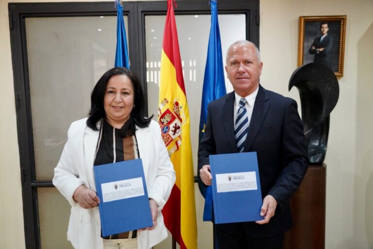 Two officials in business attire hold blue folders with an emblem, standing before Spanish and EU flags in a formal office setting.