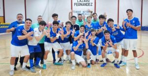 Group of young male futsal players in blue jerseys posing for a team photo in an indoor gymnasium with coaches nearby.