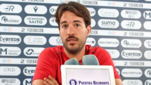 Male athlete speaking at a press conference, seated in front of a sponsor backdrop with two microphones on a tablet screen in front of him.