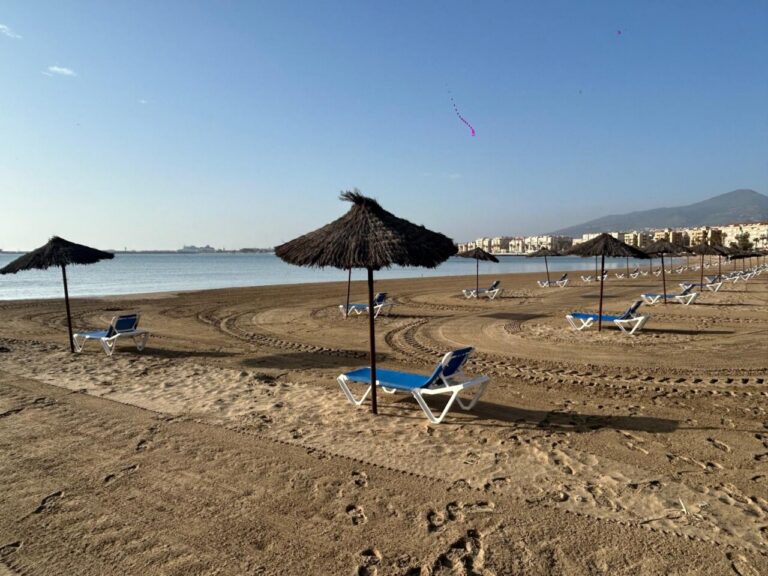Beach scene with rows of blue lounge chairs under thatched umbrellas along the shore and calm water in the background, city buildings and hills on the horizon.