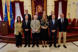 Ten formally dressed adults pose for a group photo in a ceremonial hall, with flags and a crowned crest backdrop behind them.