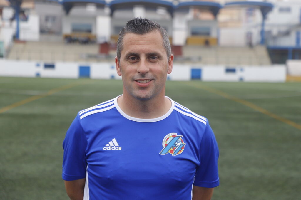 Man in a blue Adidas soccer jersey standing on a green football field with stadium seating in the background.