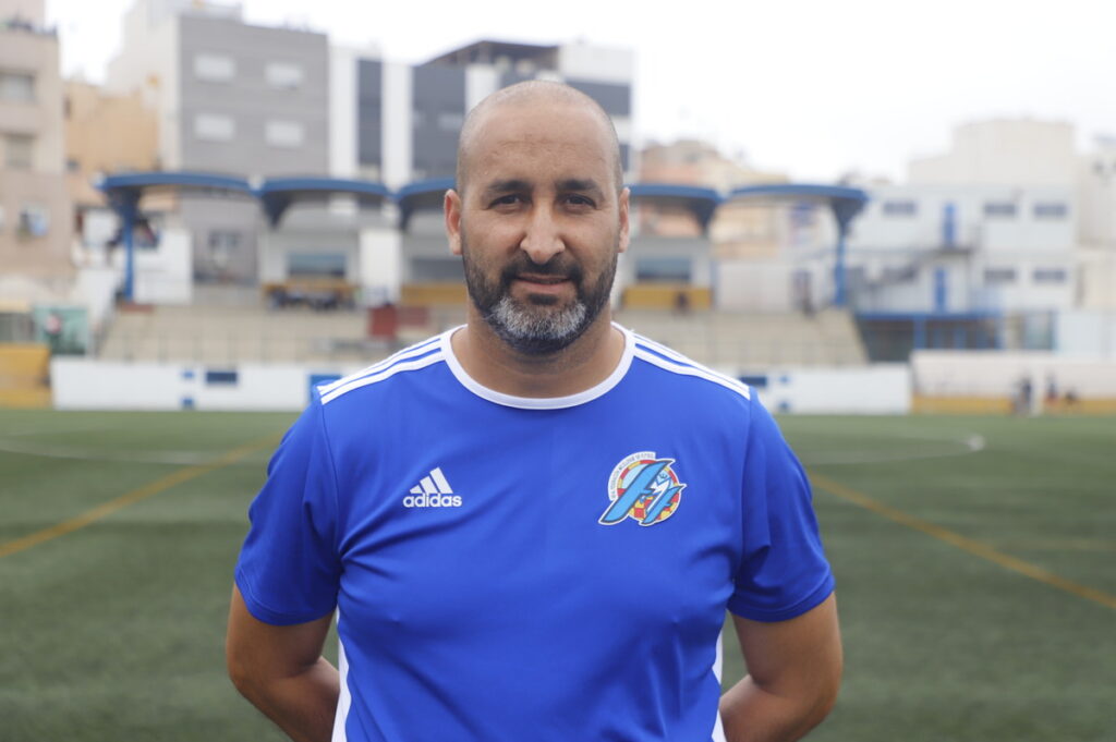Man in a blue Adidas soccer jersey stands on a blurred stadium field, facing the camera.