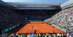 Two players on a red clay tennis court in a packed stadium, spectators all around.