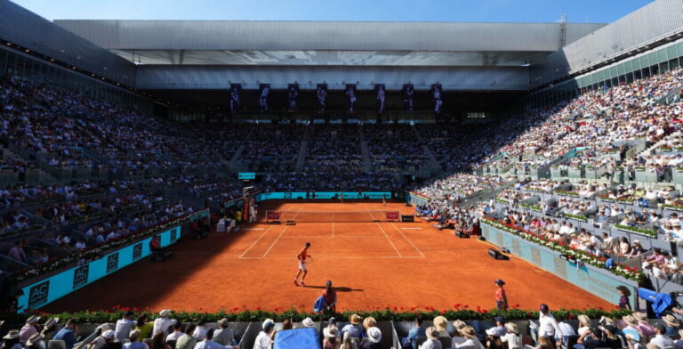 Two players on a red clay tennis court in a packed stadium, spectators all around.