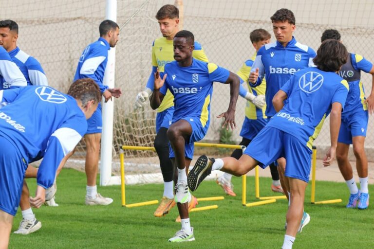 Soccer players in blue training outfits sprinting and jumping over yellow hurdles on a grass field during a practice session.