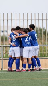 Blue-clad soccer players form a tight huddle on the field, arms around each other in celebration or strategy.