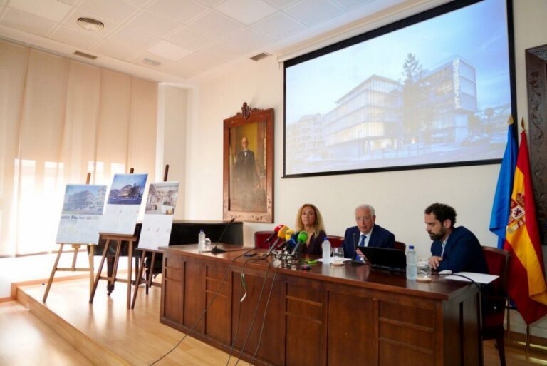 Three panelists sit at a wooden conference table with microphones, posters on easels to the left, and a large projector screen showing a modern building behind them.