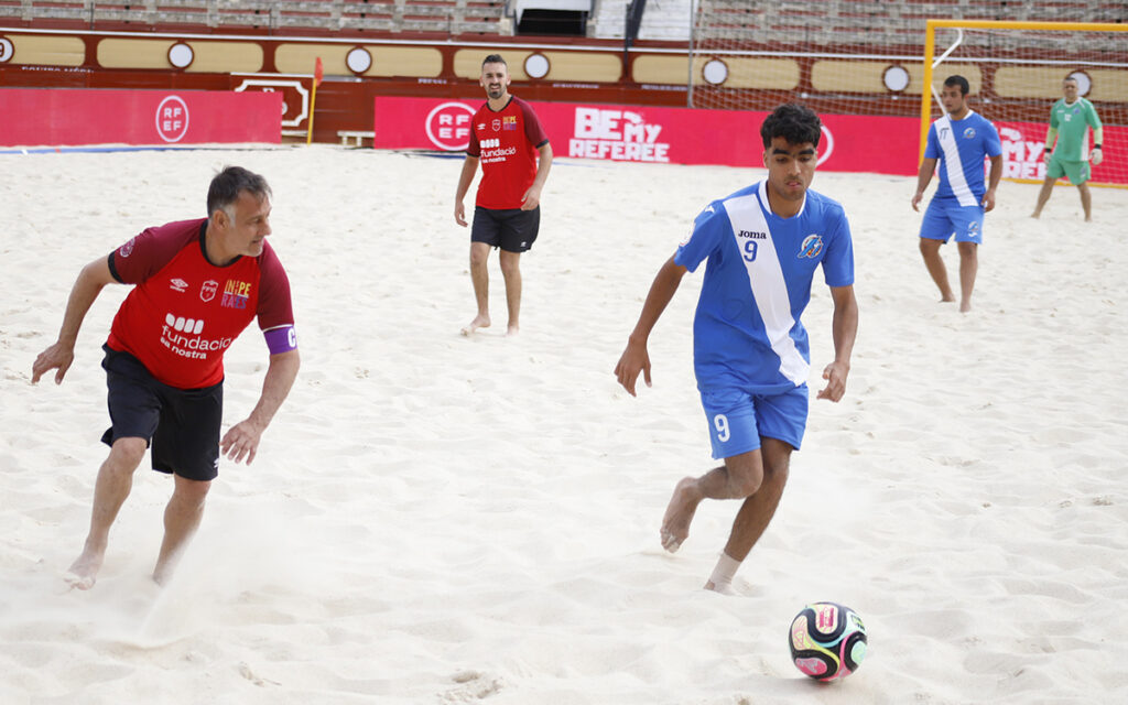 Beach soccer match: blue team player #9 dribbles the ball as a red-team opponent pursues on the sandy field.