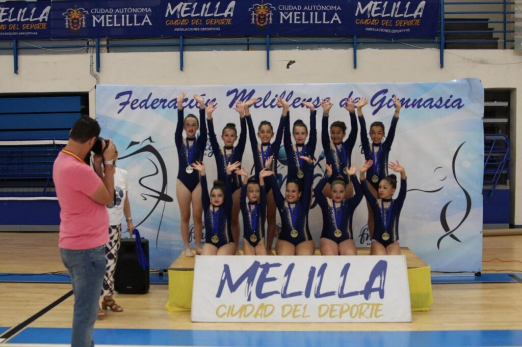 Young gymnasts pose on a podium wearing navy leotards and medals, raised hands in victory, photographer in pink shirt capturing the moment in a gym.