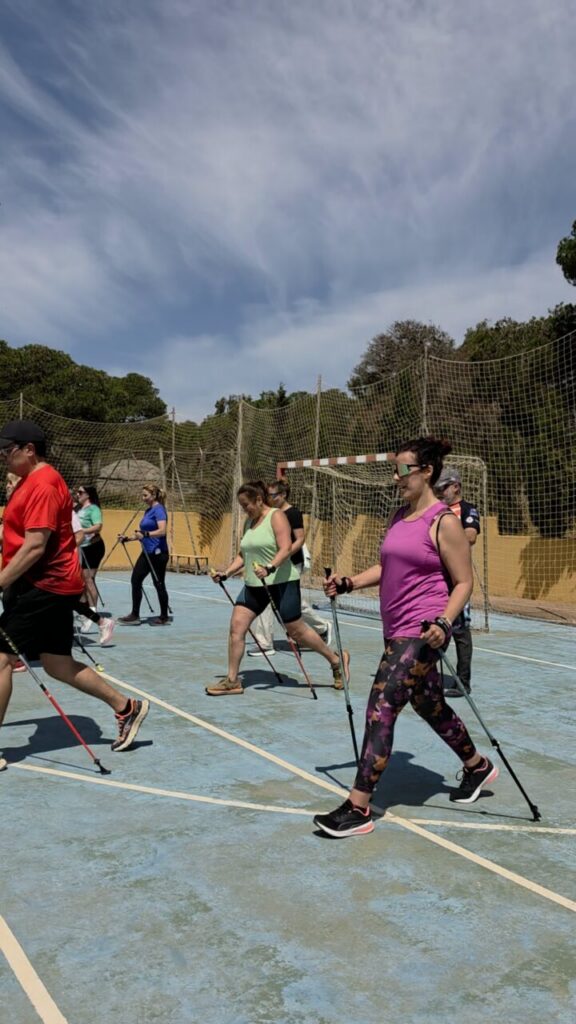 Group of people walking with Nordic walking poles on an outdoor sports court under a blue sky with a netted fence in the background.