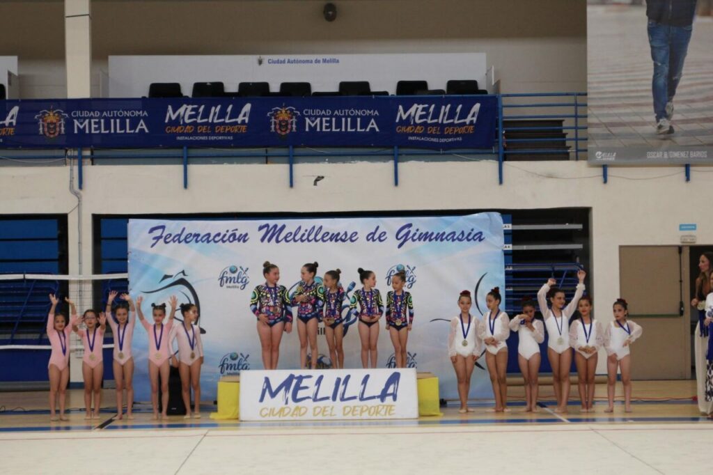 Young gymnasts in colorful leotards with medals pose on a podium at a gymnastics event, banner reads Federación Melillense de Gimnasia in the background.
