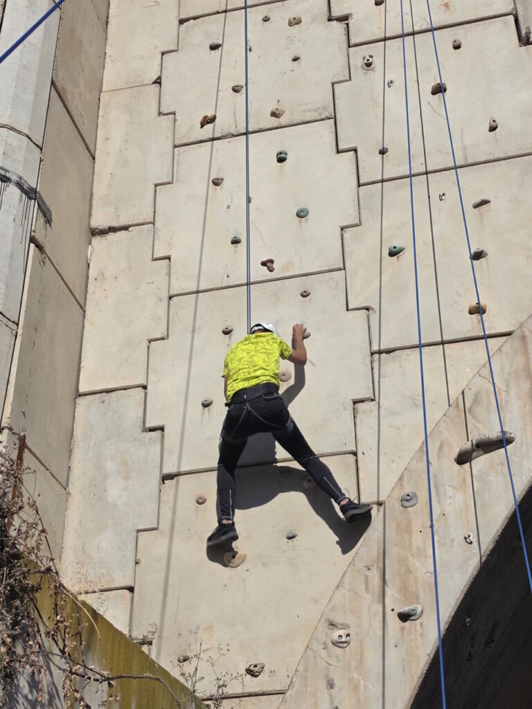 Person wearing a yellow shirt and helmet climbs an outdoor artificial rock wall with blue ropes for safety.