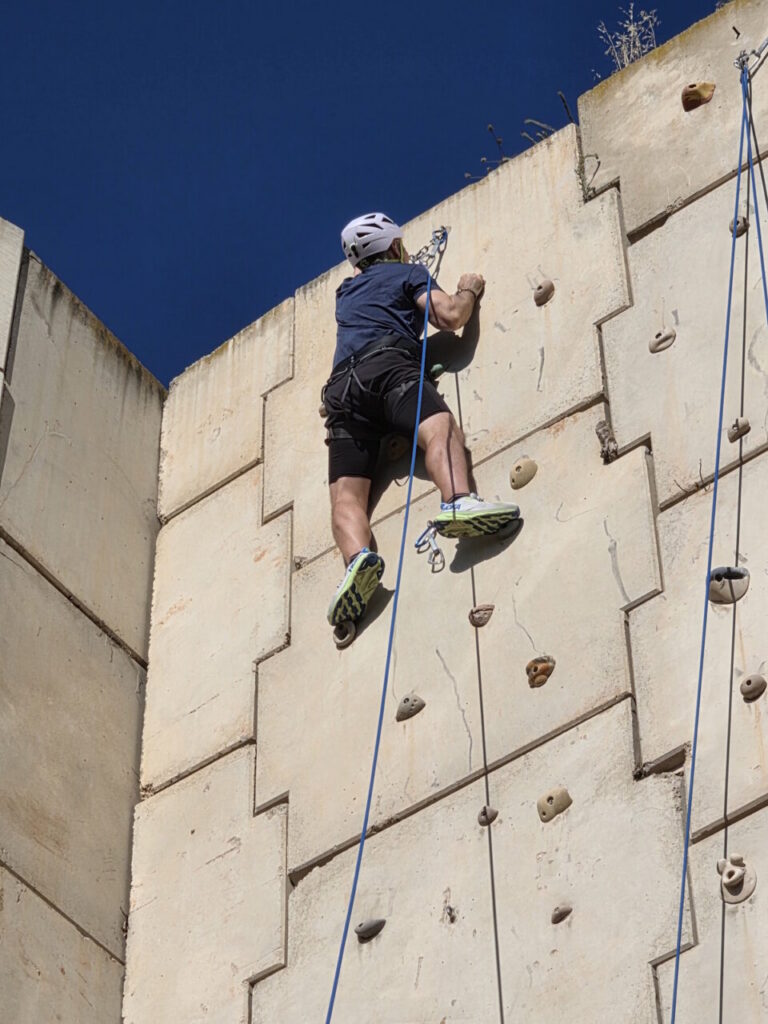 Climber wearing a helmet and harness ascends a tall outdoor rock wall using ropes against a blue sky.