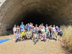 Group of people in athletic wear posing in front of a large tunnel opening, outdoor dirt ground; one person in a handcycle wheelchair at center.