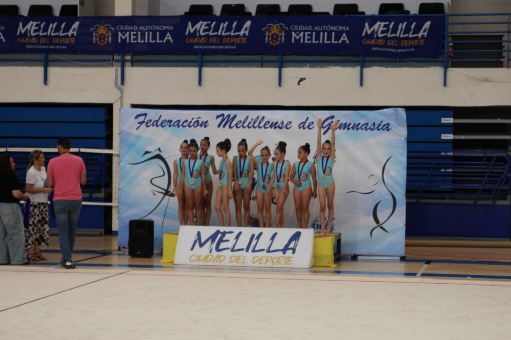 Young gymnasts in turquoise leotards stand on a podium with medals during a ceremony in a gymnasium with banners overhead.