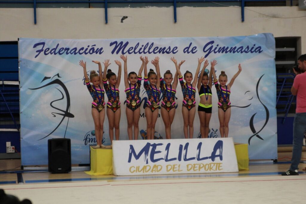Group of young female gymnasts on a winner's podium with arms raised in celebration, wearing colorful leotards and medals, in front of a banner that reads Federación Melillense de Gimnasia.