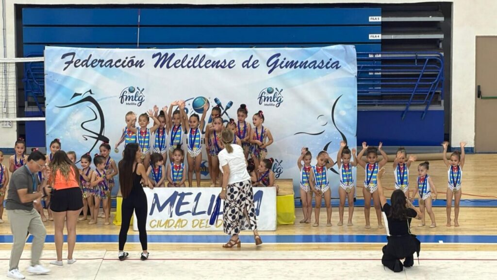 Young gymnasts on a podium with medals during an awards ceremony in a gym, with a banner reading Federación Melillense de Gimnasia.