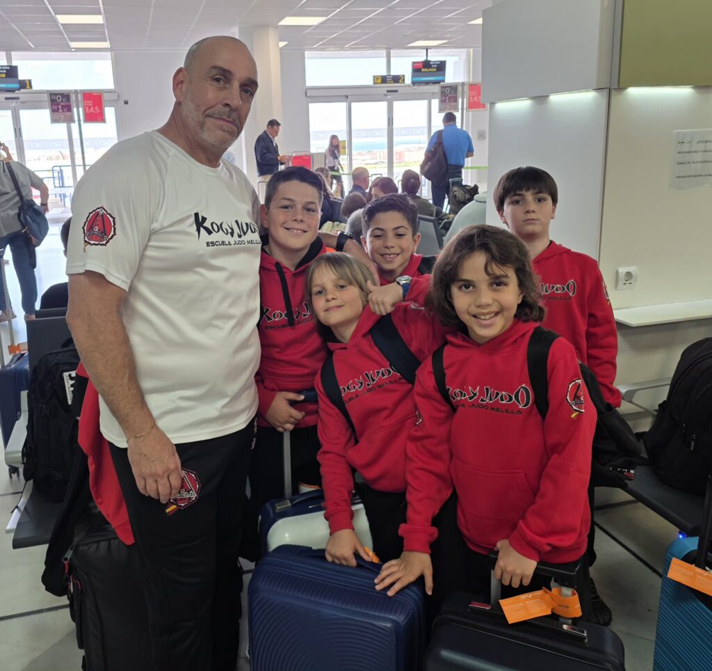 Group of smiling children in red hoodies posing with a man in a white T-shirt at an airport check-in area with suitcases nearby.