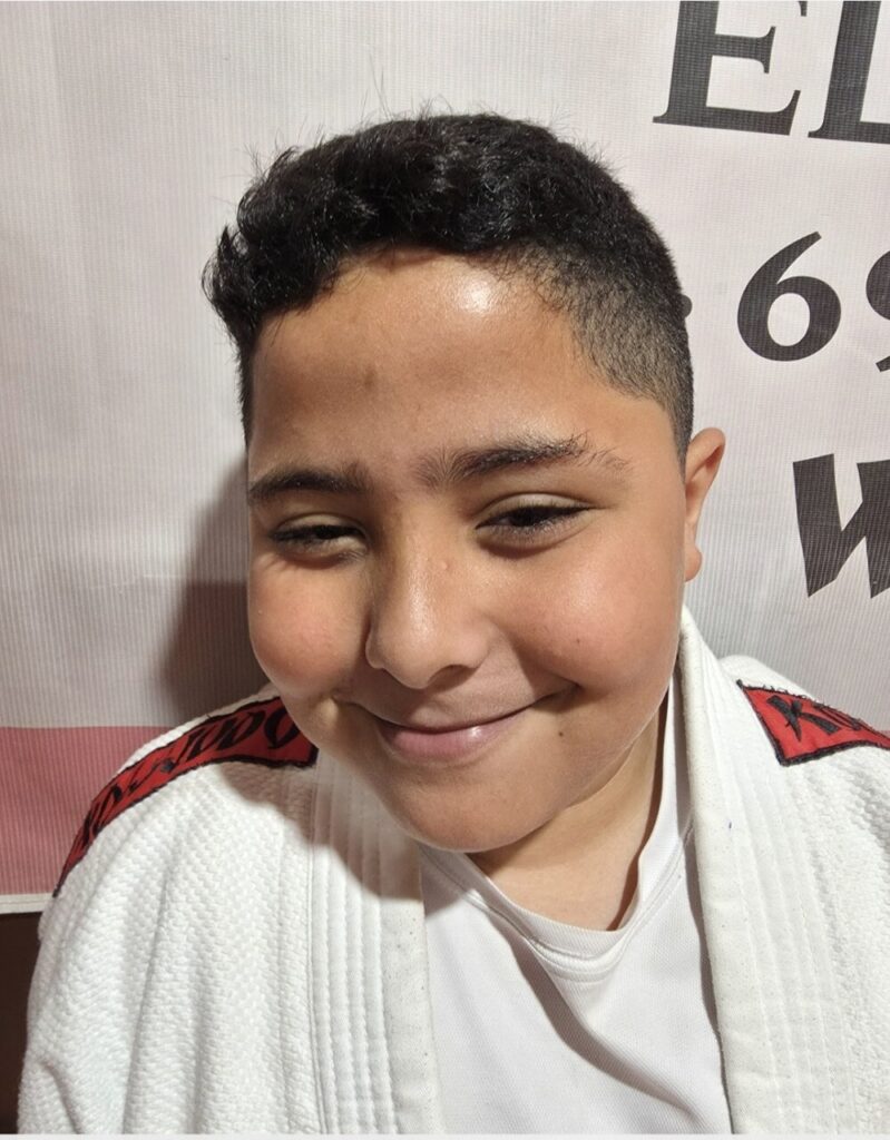 Smiling young person wearing a white martial arts gi with red patches on the shoulders, posing for a photo indoors.