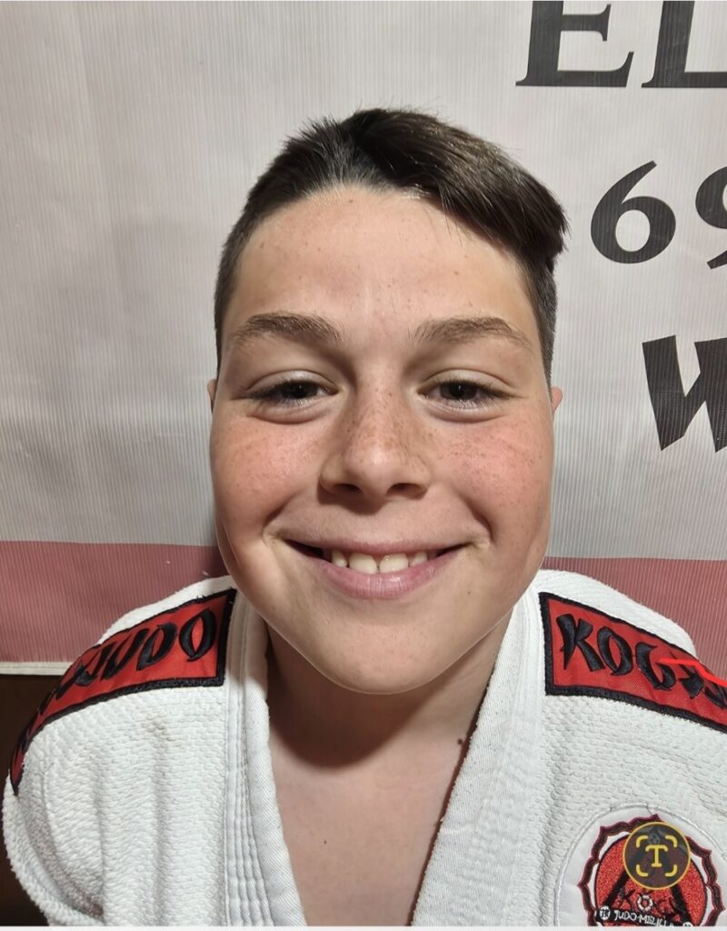 Smiling child in a white judo gi with red shoulder patches, standing in front of a backdrop with text and logos on the right shoulder badge visible.