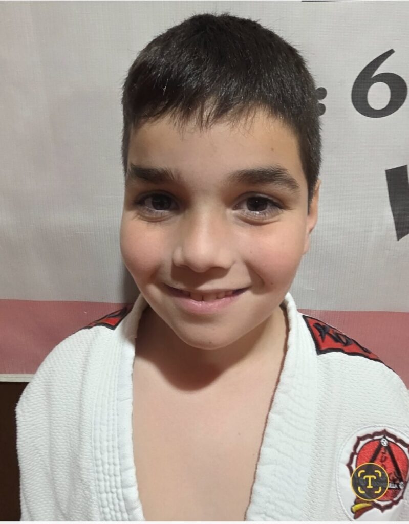 Young boy with short dark hair smiling at the camera, wearing a white martial arts gi with a red patch on the shoulder.