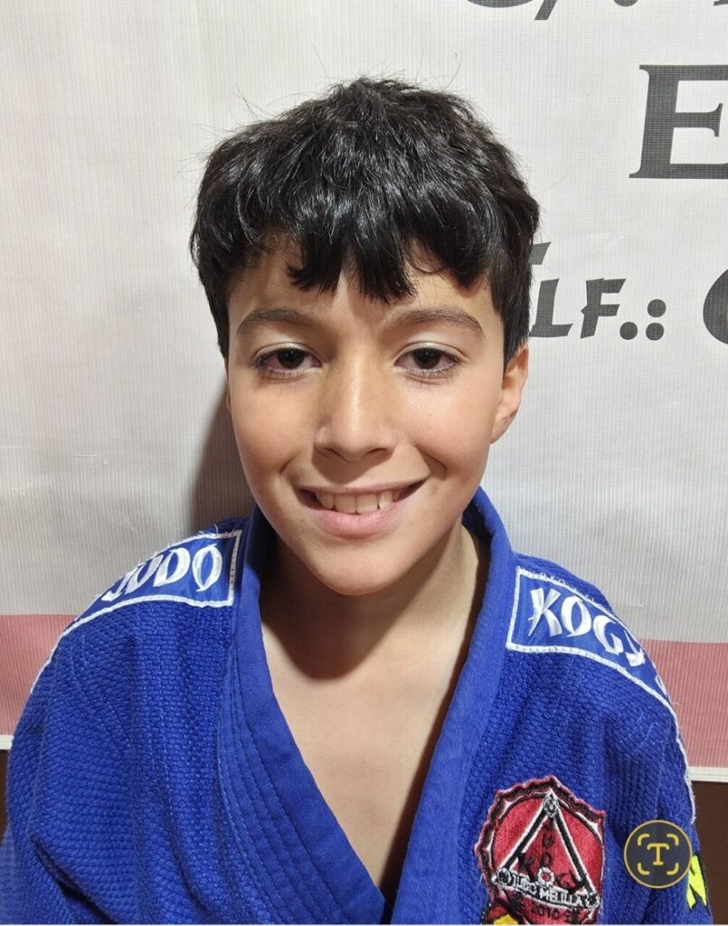 Smiling boy in a blue martial arts gi with patches, posing for a portrait in a dojo setting