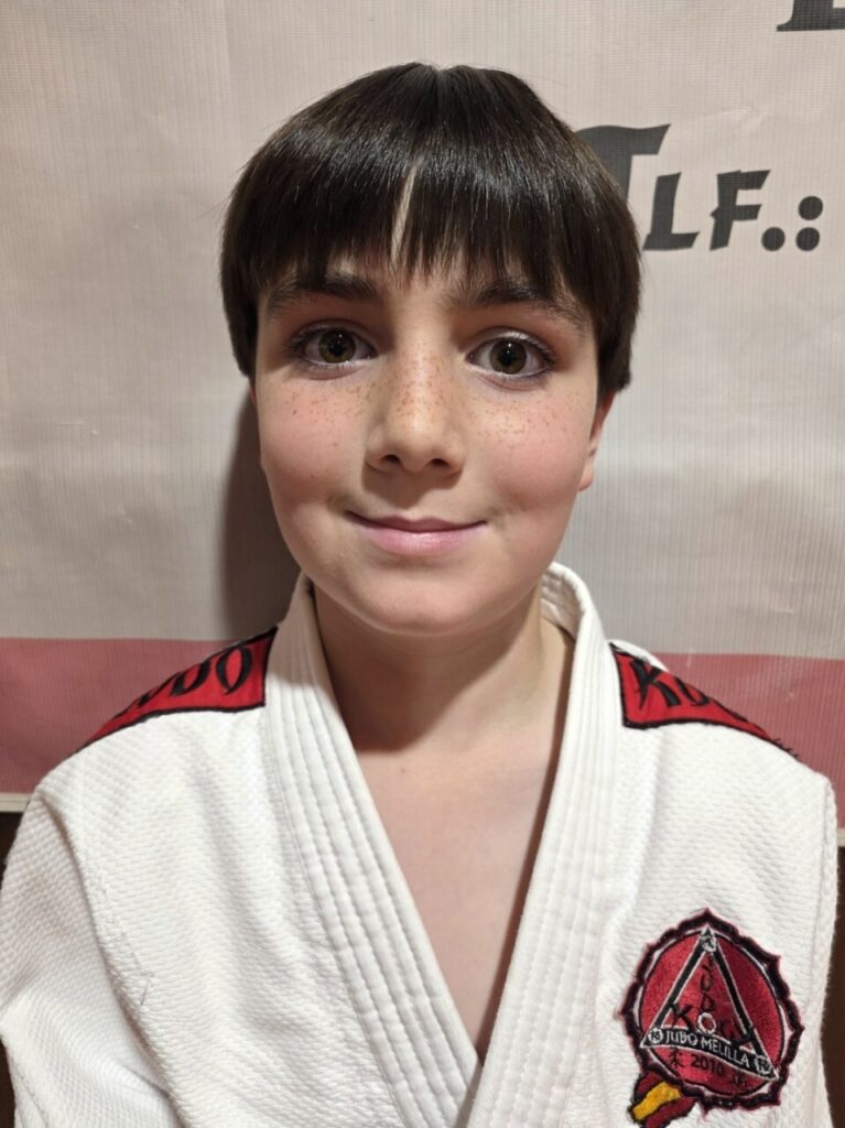 Close-up of a smiling boy with a dark bowl haircut and freckles, wearing a white martial arts gi with red patches and a crest on the chest.