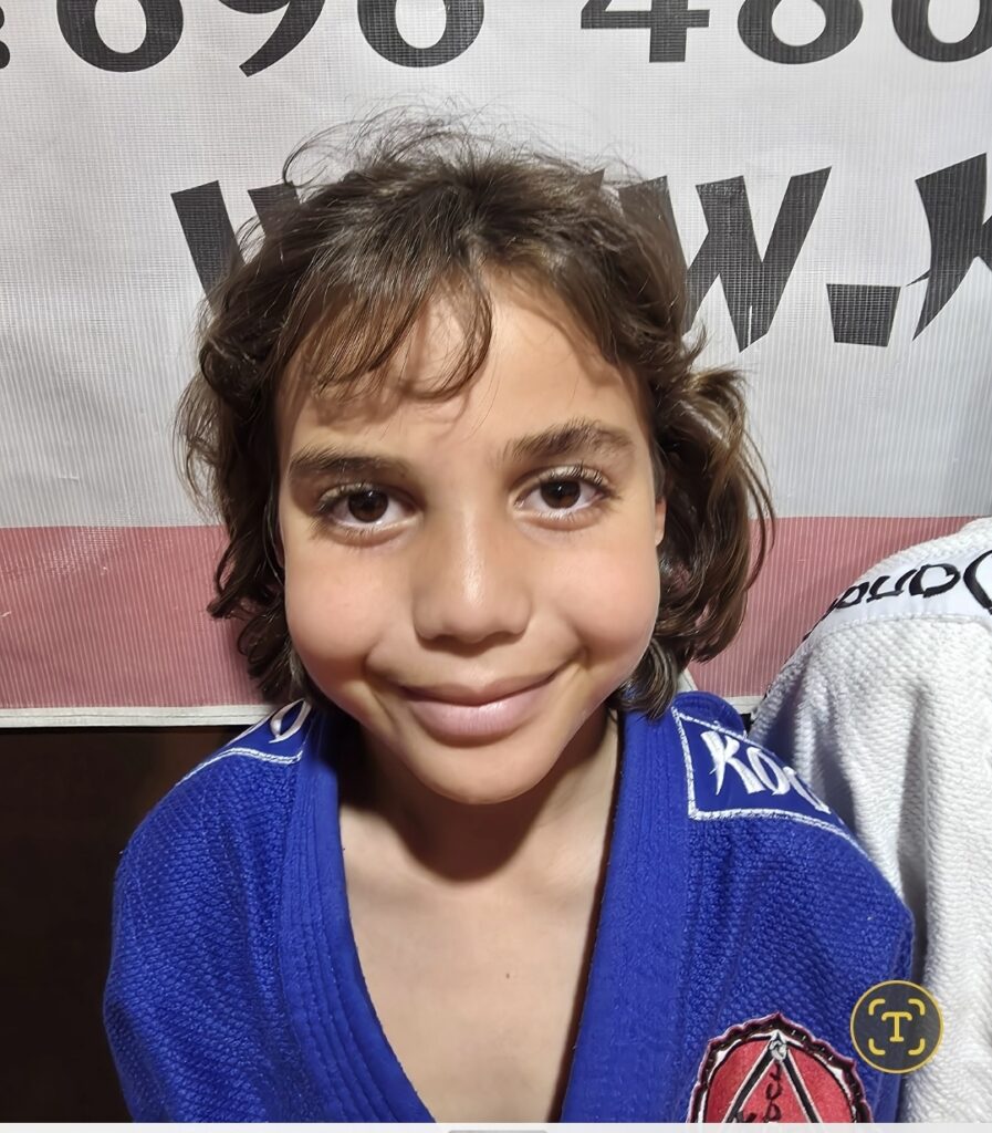 Smiling young child with brown hair wearing a blue judo gi with patches, standing in front of a banner with large numbers.