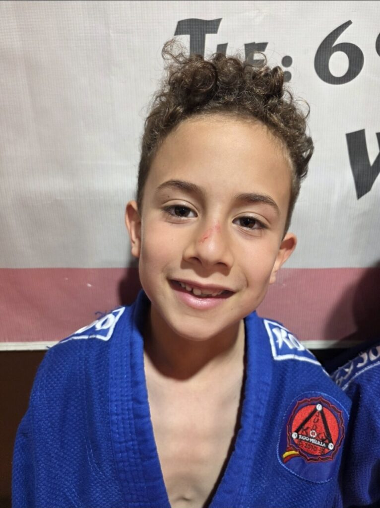 Smiling young boy with curly hair in a blue martial arts gi, standing in front of a banner.