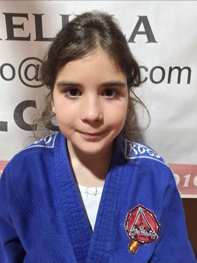 Young girl in a blue judo gi smiling at the camera, with a red shield patch on the left chest and a banner backdrop behind her.